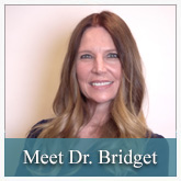 The image shows a woman with long hair, wearing a dark top, posing for a portrait with a professional backdrop. She has a friendly expression and is smiling at the camera. The background includes text that reads MEET DR. BRIDGET.