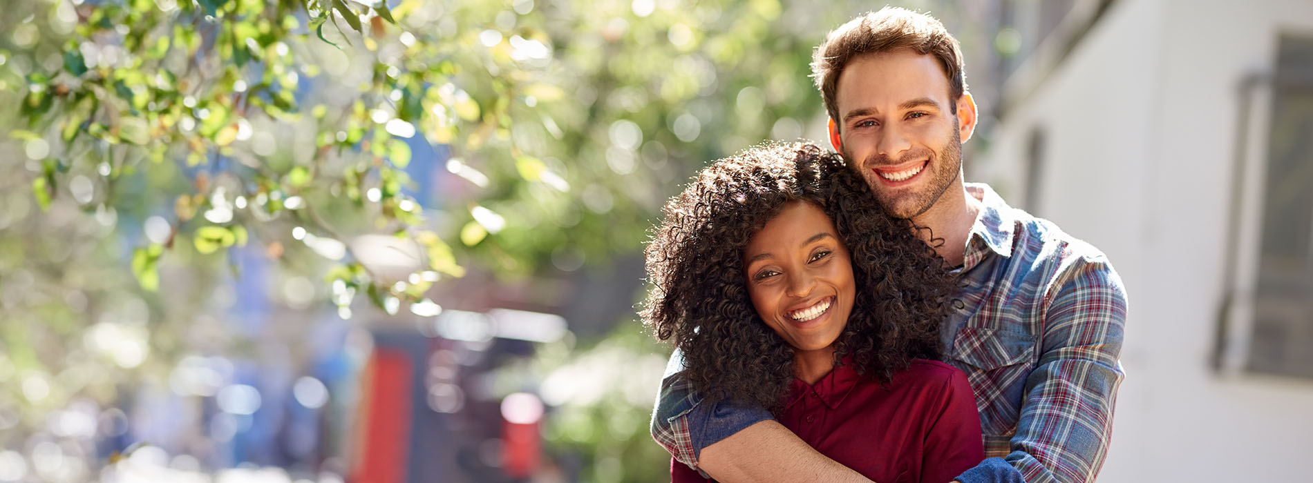 A man and woman are hugging each other outdoors the man is wearing a plaid shirt and has short hair, while the woman is wearing a dark top with a lighter sleeve and has long hair.